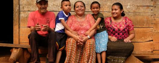 Aurelia and her family sit on a wooden bench outside their home, smiling. She holds a young boy while a little girl hugs her. A man in a red t-shirt and a woman in a pink blouse smile beside them.